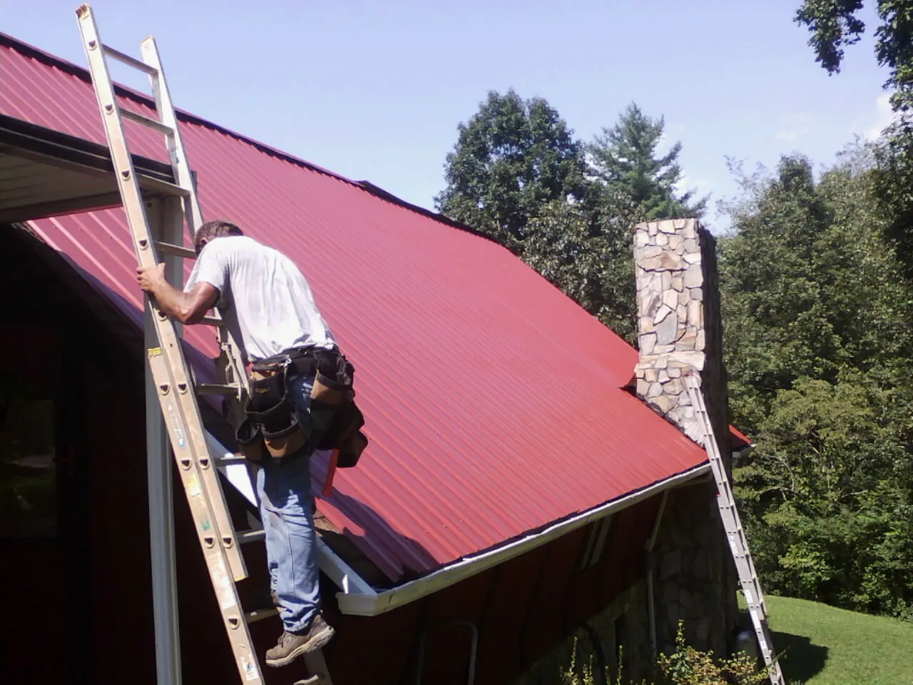 Roofer on a ladder beside a red metal roof and stone chimney — Soffit & Fascia Repair by Roofing Brothers in Richburg